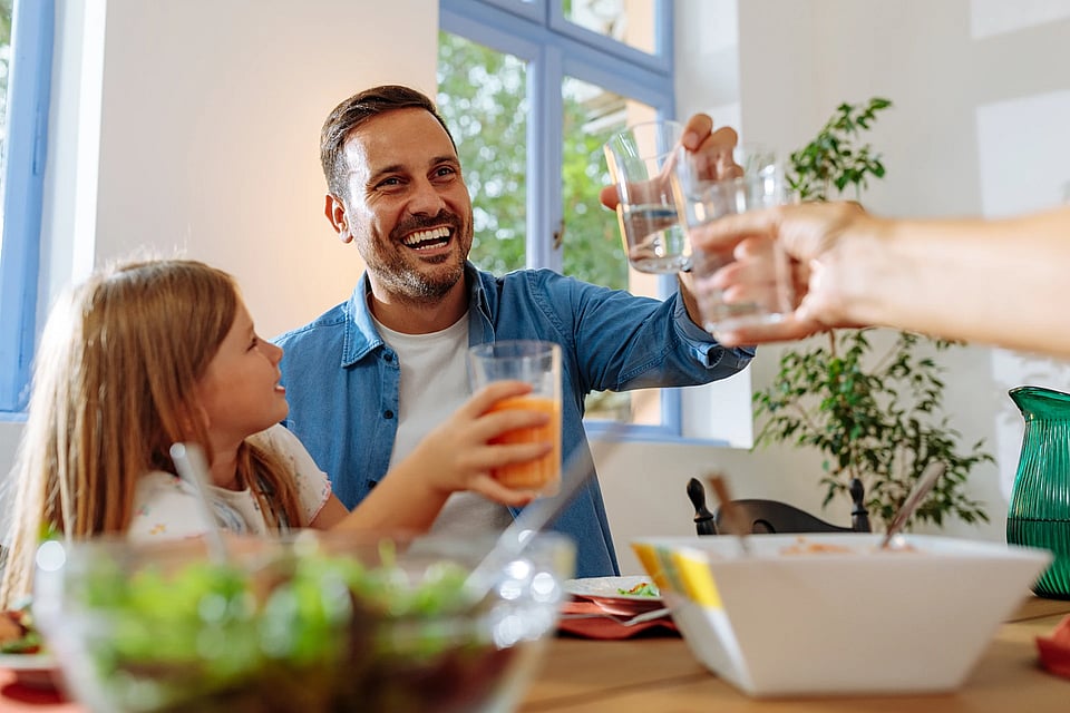 A dad and daughter clinking water glasses at the dinner table and smiling.