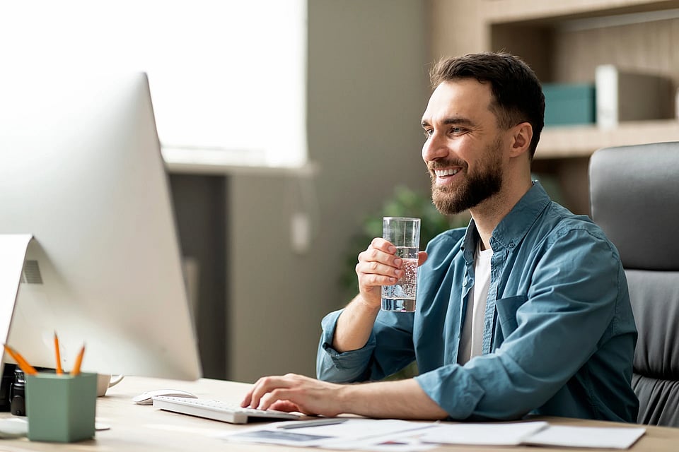 A man working at a computer in an office while enjoying a glass of water.