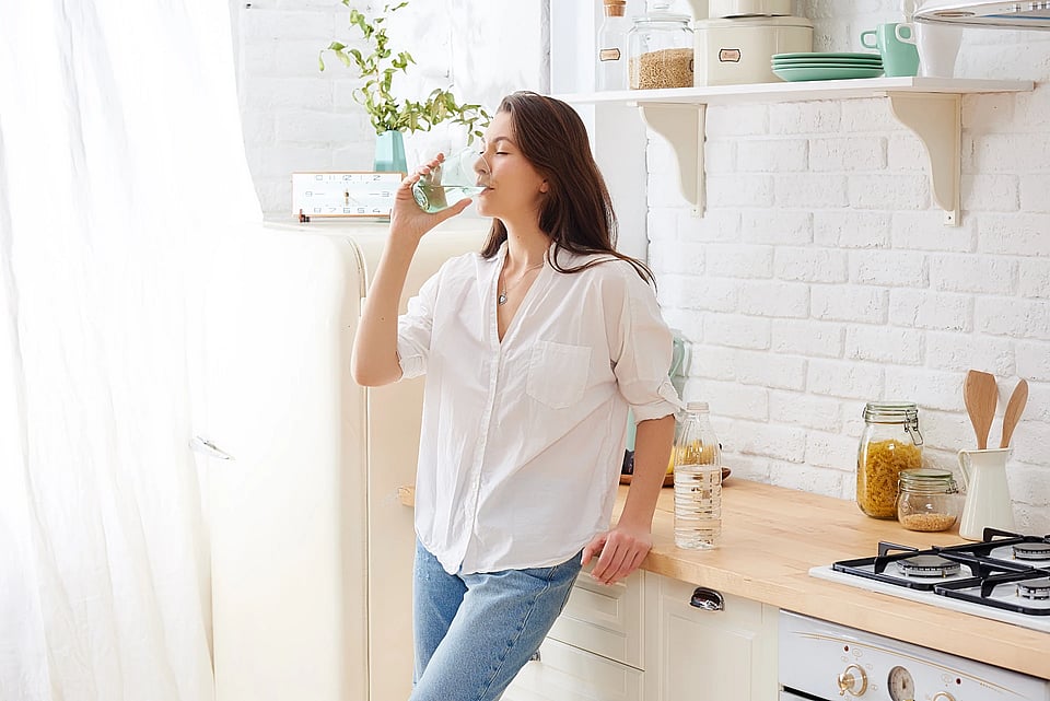 A woman in a white shirt and jeans, standing by her counter enjoying a glass of clear water.