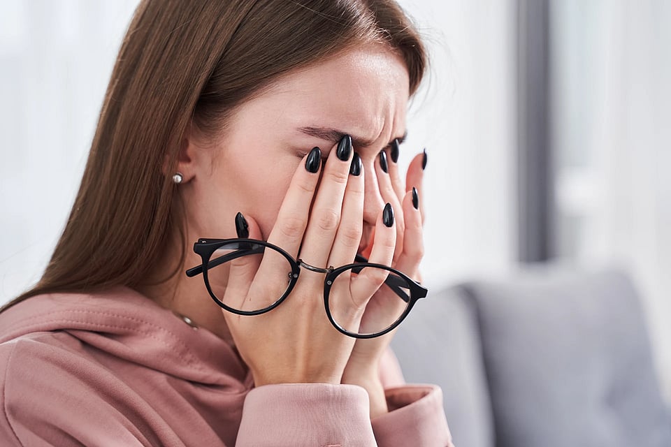 A woman with brown hair and black nails holding black glasses and rubbing her eyes like they're bothering her.