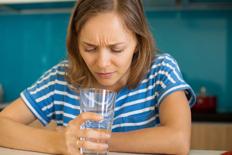A woman in a blue shirt sniffing a glass of water that smells bad.