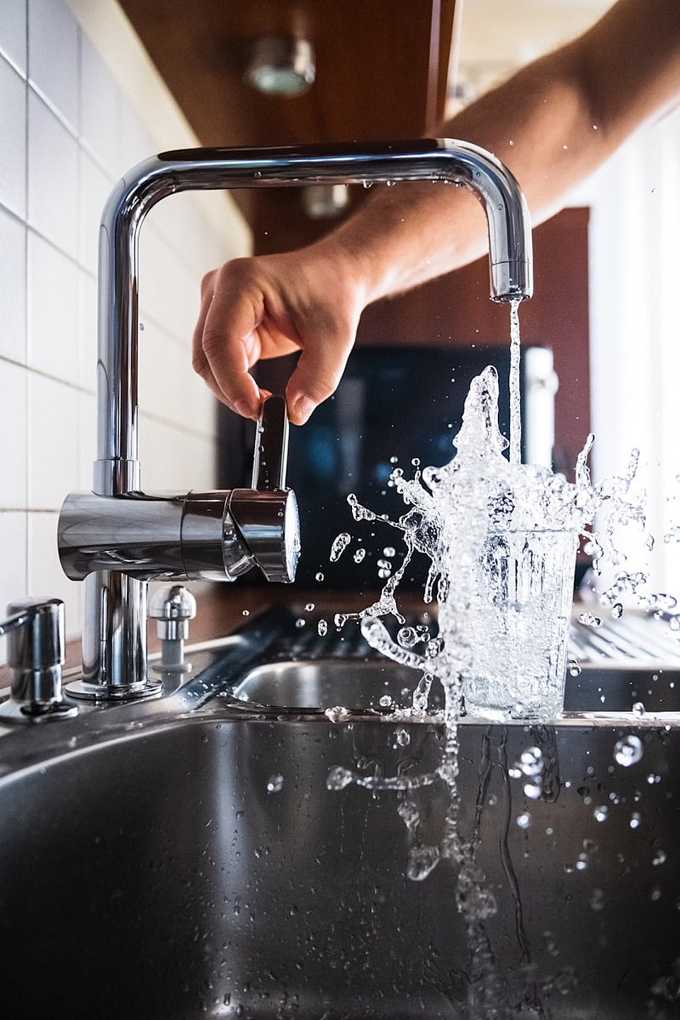 Hand turning a kitchen faucet as water splashes into a drinking glass