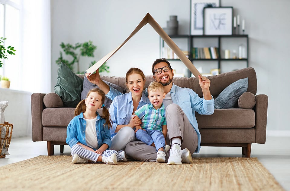 A family by a couch, holding a piece of cardboard over their heads like the roof of a house