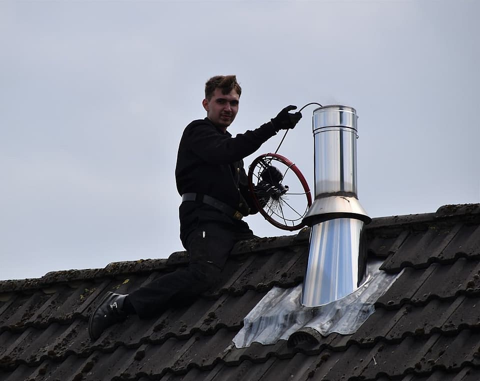 Chimney sweep technician on a tiled roof holding a sweeping brush beside a stainless steel chimney flue