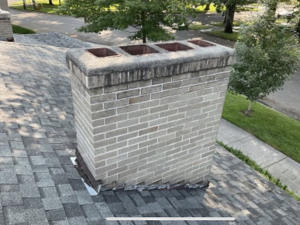 Tan brick chimney with a concrete crown and four clay flue liners on a shingled rooftop